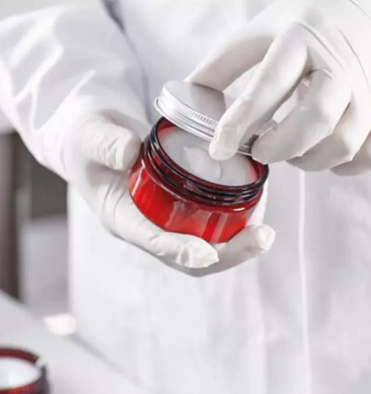 A gloved hand checking cream in a glass jar in the lab